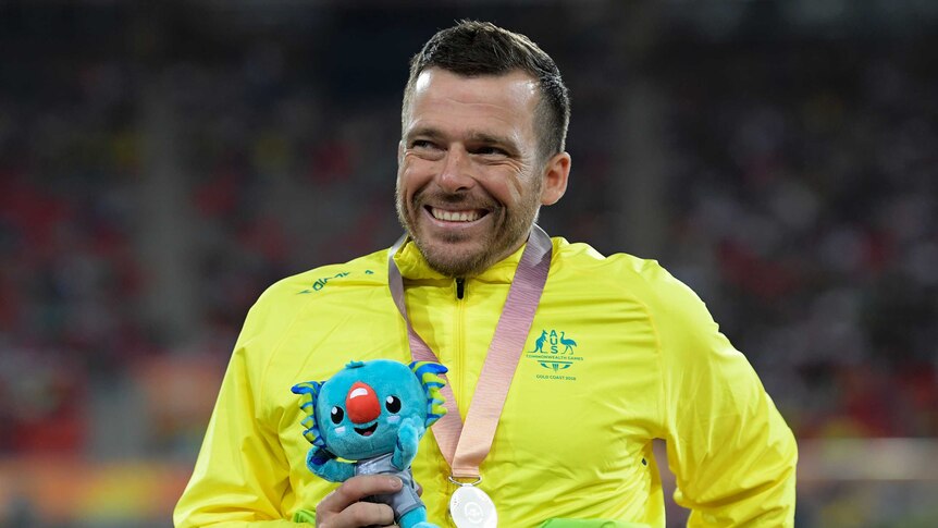 Kurt Fearnley smiles with a silver medal around his neck and a Commonwealth Games mascot in his hand.