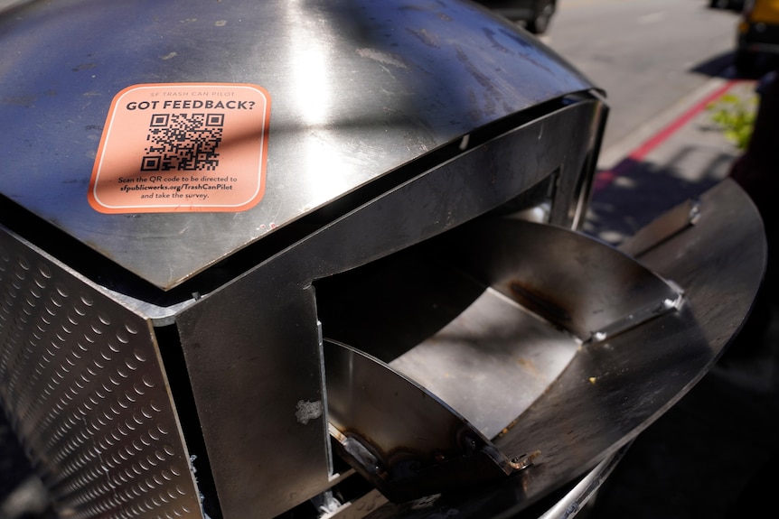 A close-up of a beige QR code sticker, with the words "GOT FEEDBACK?", on top of a smooth metal garbage bin.