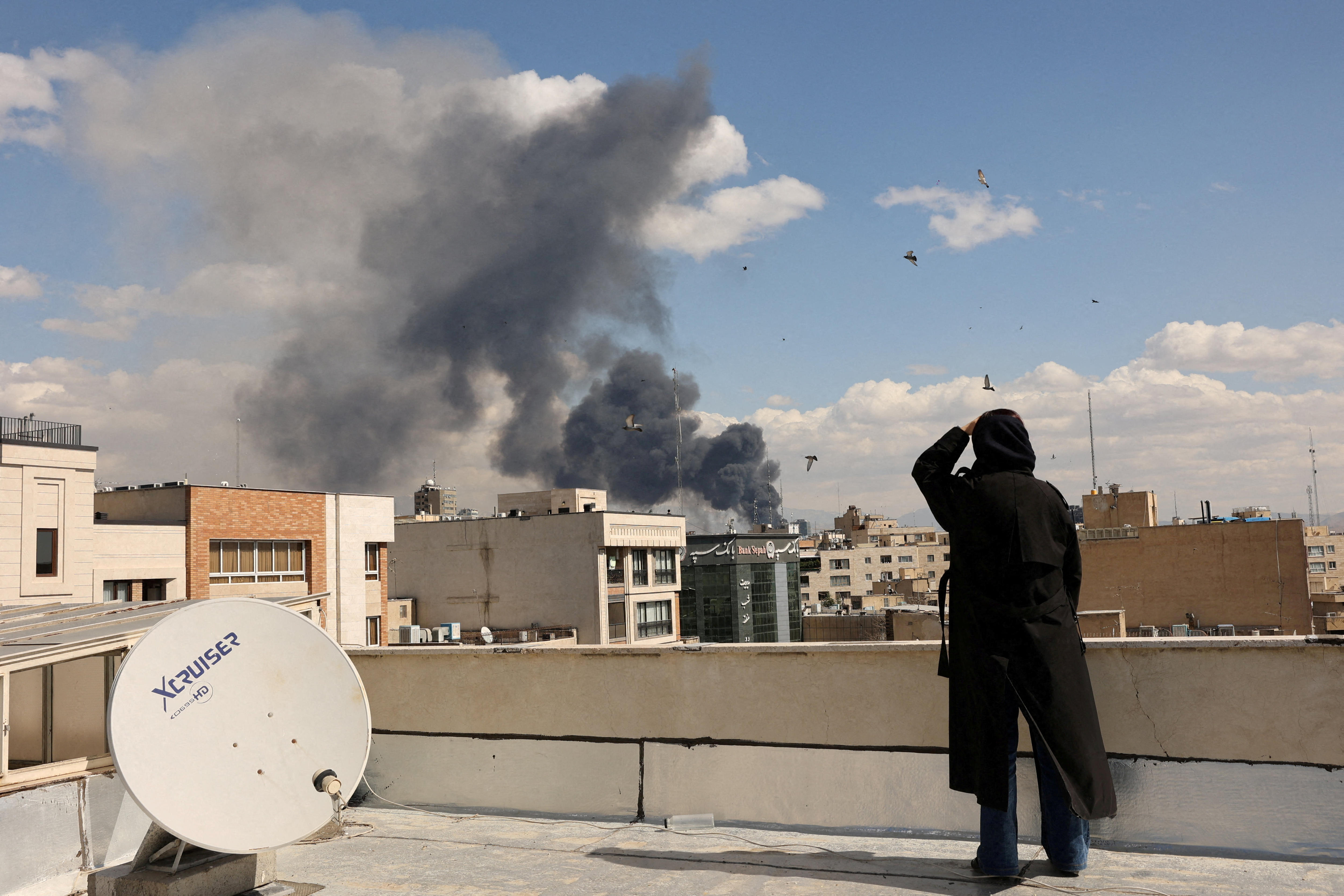 Una persona vestida de negro mira desde un tejado una nube de humo que se eleva desde un edificio a lo lejos. 