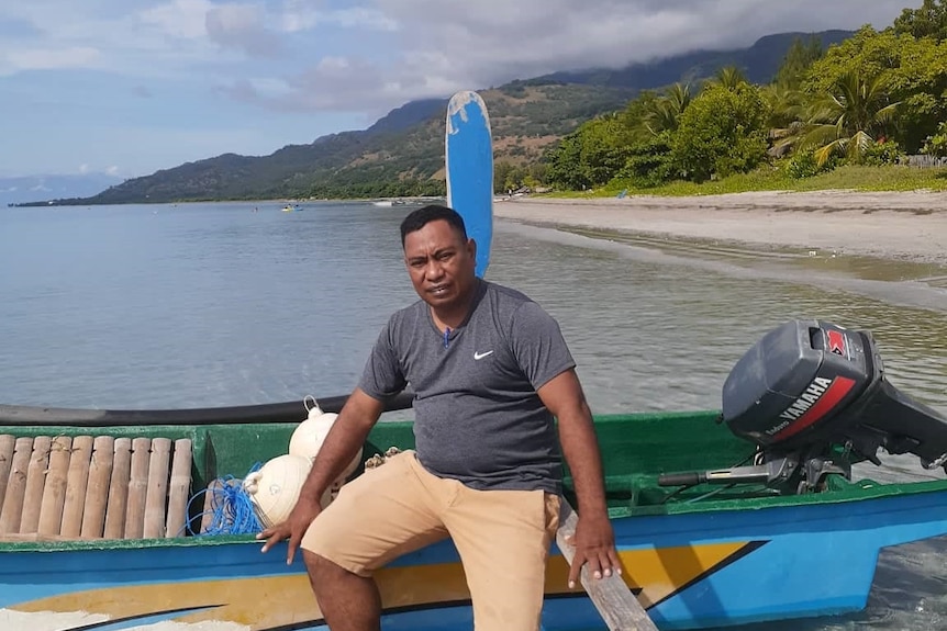 A man sits on the sideof a boat with a beach and trees behind him