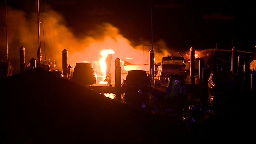 Firefighters hold hoses towards a boat which is well alight on a marina.