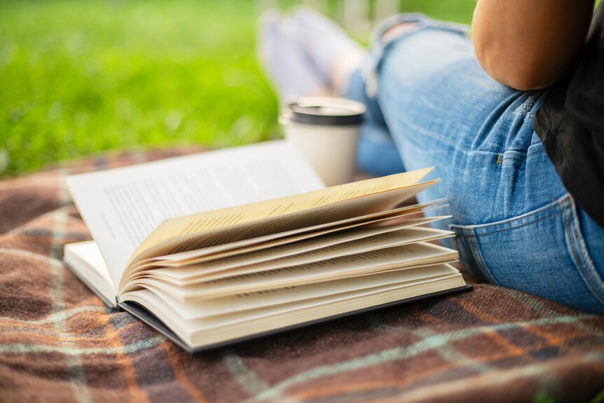 An open book sits on a brown plaid picnic rug beside a person wearing jeans who has a takeaway coffee cup.
