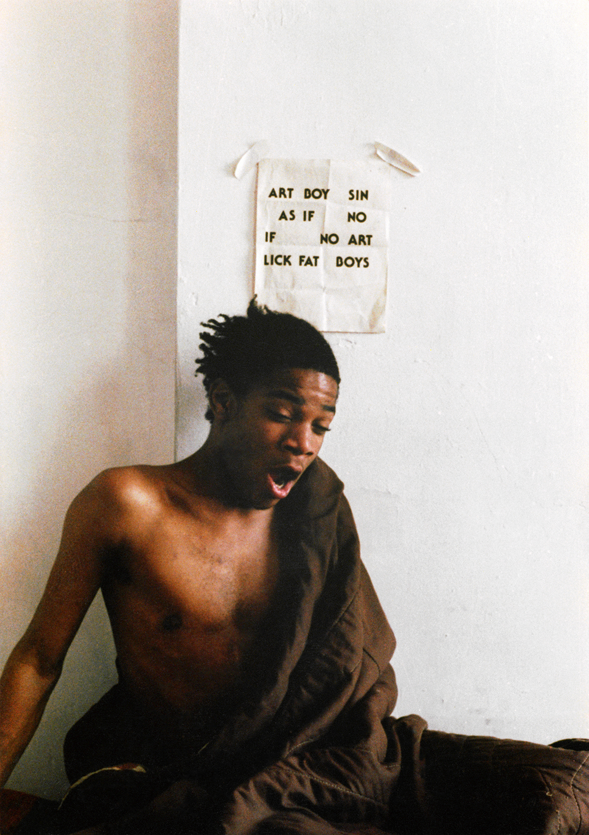 Colour photograph of Jean-Michel Basquiat sitting under a paper sign on a white wall and yawning, draped in a brown fabric.