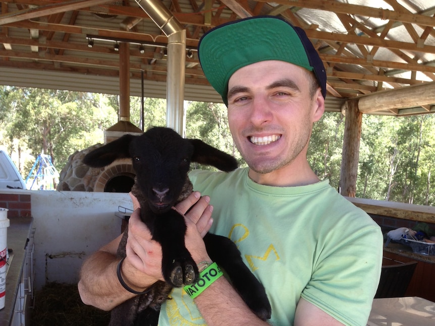 Man in green t-shirt smiles holding a small black goat. 