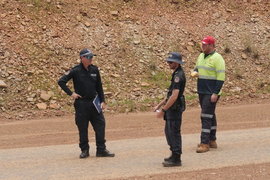 Two police officers and a man in high-vis stand on a remote road 