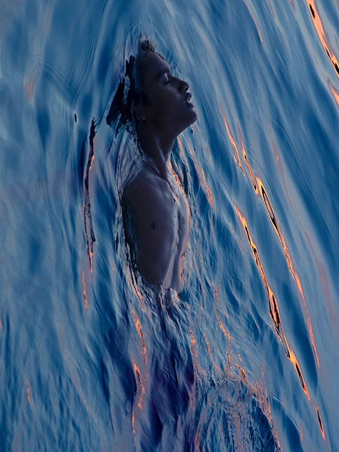 A boy with eyes closed floats in the ocean at dusk or dawn, only his face and top half of chest are above the surface.