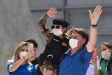 Jair Bolsonaro holds a child in military dress waving to a crowd. All wearing face masks.
