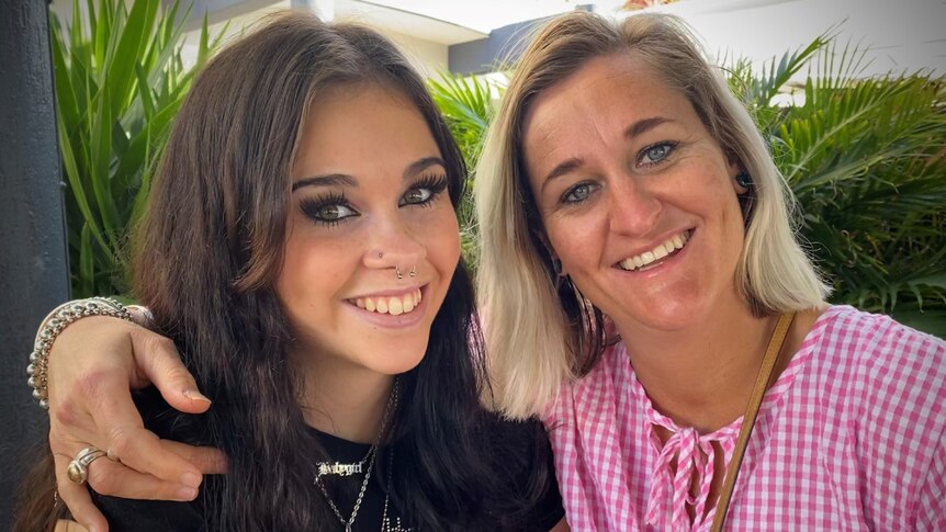 Teenage daughter with long, dark hair sits closely with mum who has blonde hair, both smiling.