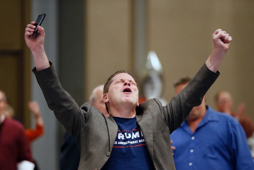 A man wearing a Trump T-shirt raises his fists in the air.