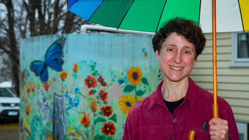 A woman holding a colourful umbrella smiles to camera, a water tank painted with flowers and a butterfly behind.