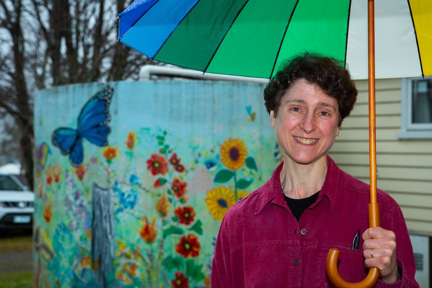 A woman holding a colourful umbrella smiles to camera, a water tank painted with flowers and a butterfly behind.