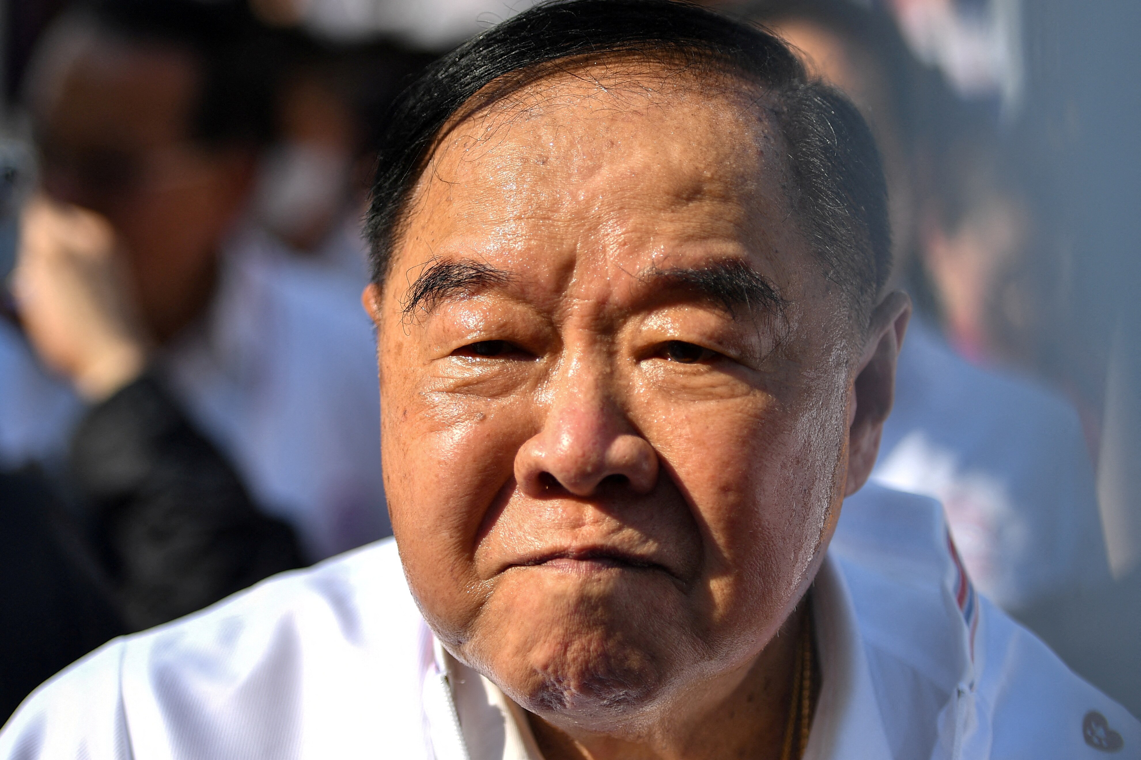 A man with short black hair combed over to one side looks sternly at the camera while lightly sweating