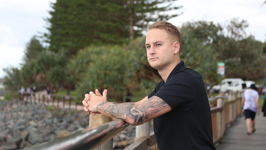 A man leans on a silver rail on a beach-side timber boardwalk with pine trees in the background.