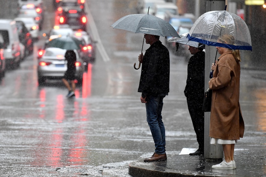 people holding umbrellas while waiting on the footpath to cross the street on a rainy day