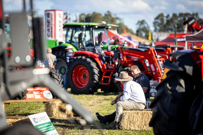 Two men sit on a hay bale with a red tractor in the background.