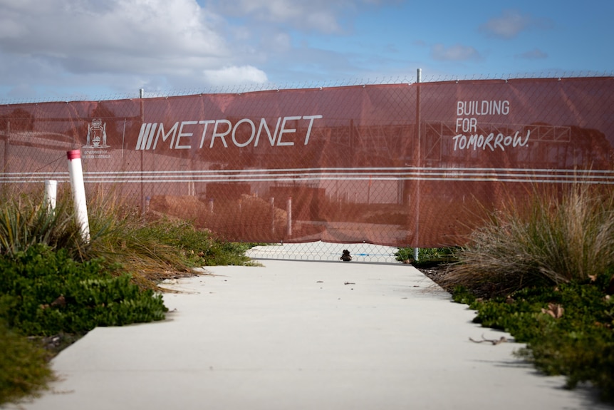 A fence with "Metronet, Building for Tomorrow" on it, crossing through the middle of a footpath.