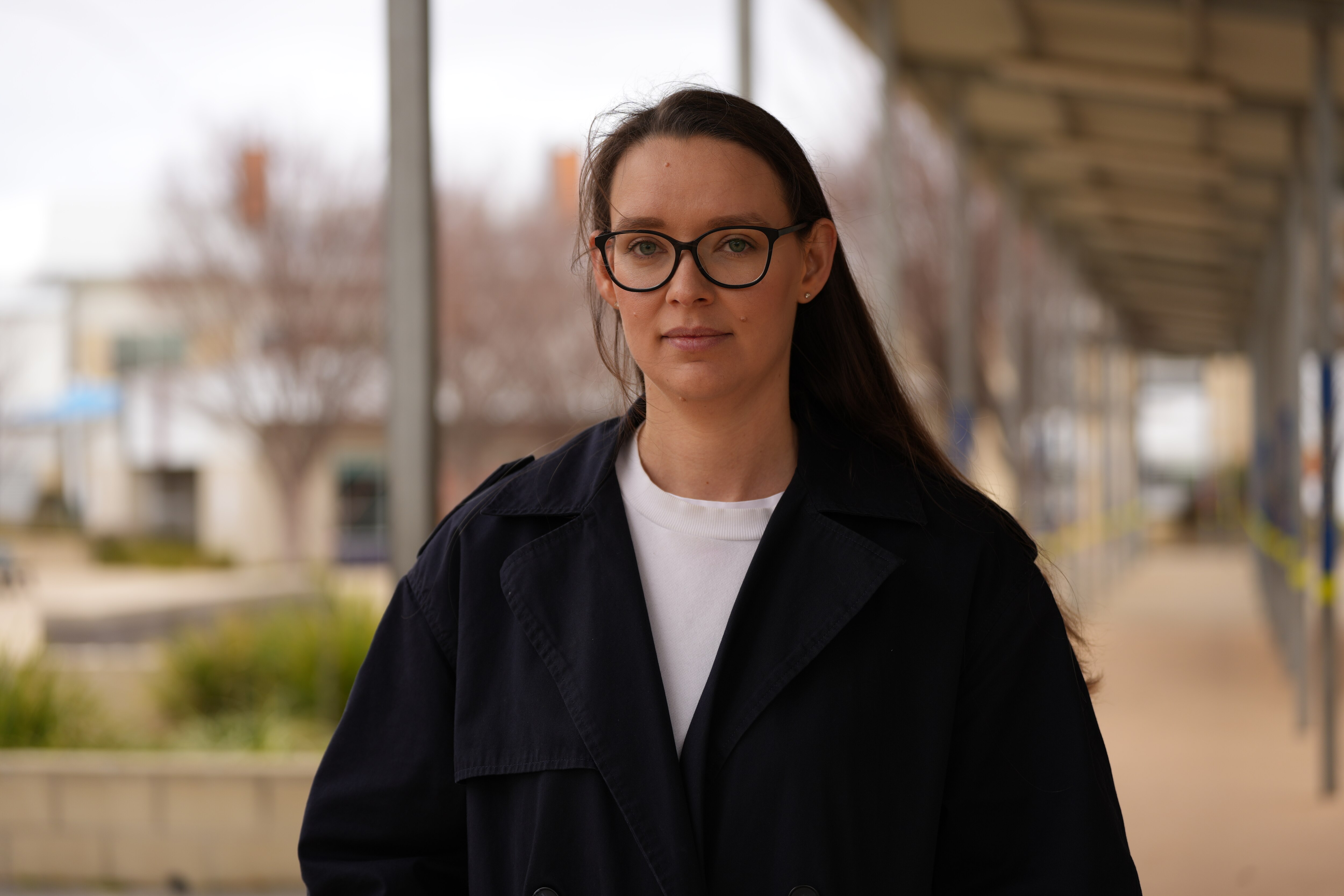 A woman with long dark hair and glasses stands outdoors looking serious.