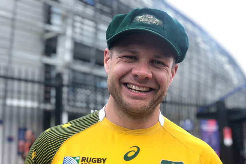 A man smiles and wears a gold jersey and a green hat