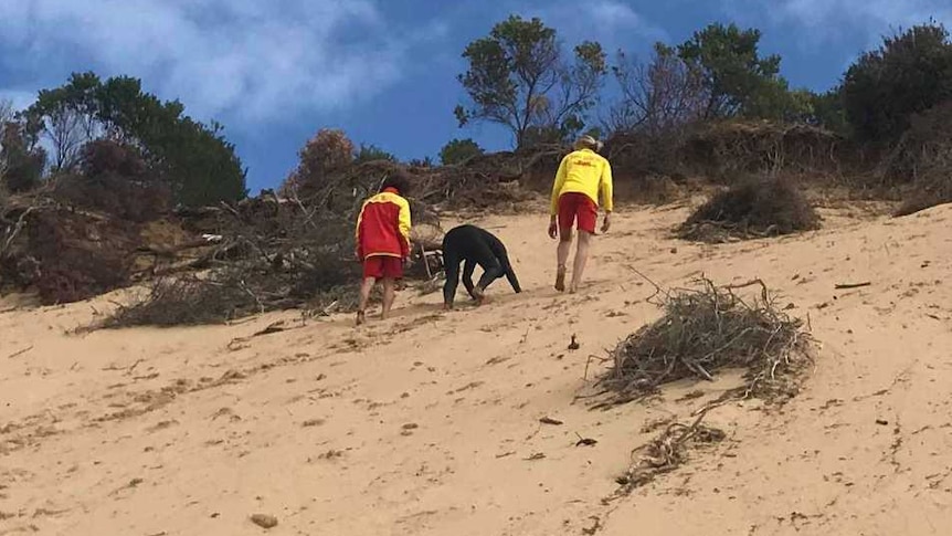 Three people in wetsuits and surf-lifesaving gear scale a steep sandhill at the beach.