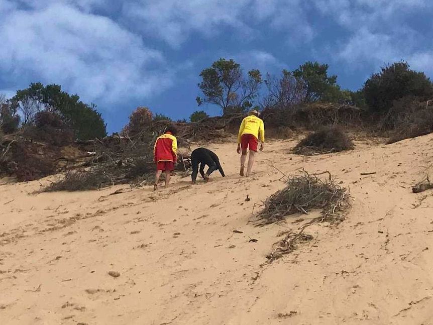 Three people in wetsuits and surf-lifesaving gear scale a steep sandhill at the beach.