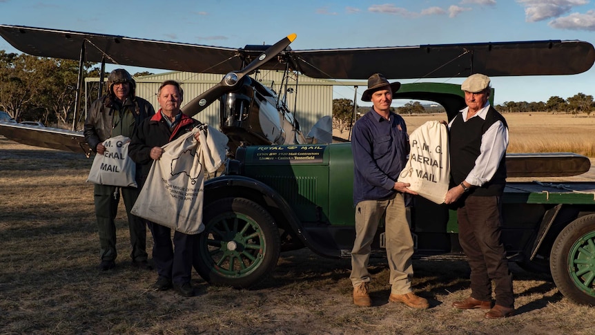 Four men stand in front of a gypsy moth biplane holding calico sacks with P.M.G Air Mail written on them.