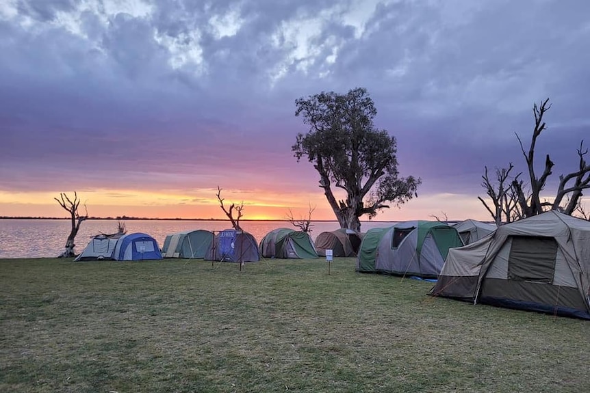 Campsites set up by the river, near some trees. The sky is cloudy but the sunrise is orange and yellow. It's early morning.