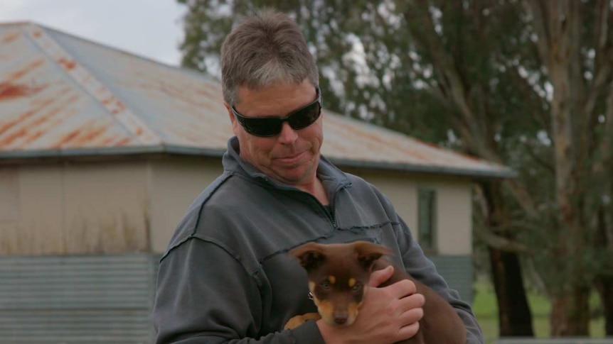 A man holding a kelpie puppy in front of a rural property