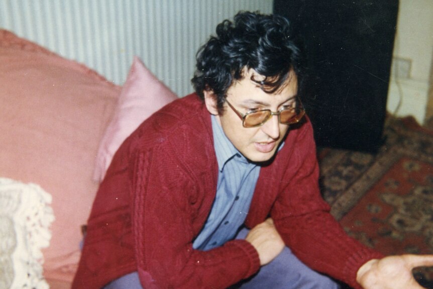 University student Rai sitting in a red cardigan and glasses. He gestures mid-conversation with one hand resting in his lap.