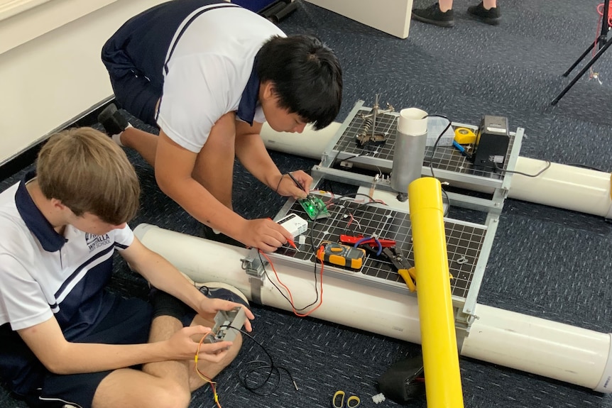 Two boys are seated on the ground inspecting and soldering components for a floating ocean rig.