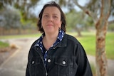 A woman stands in a park wearing a dark denim jacket with a faint smile.