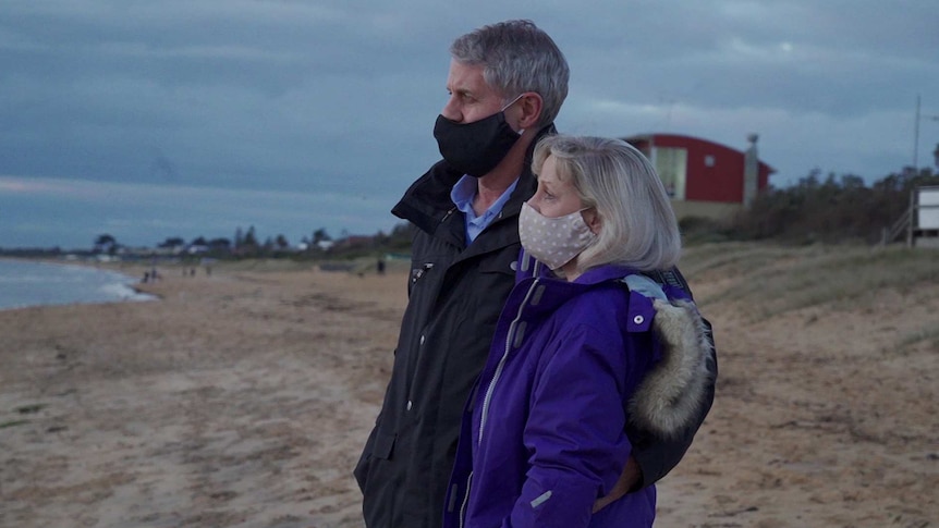 A man and woman both wear face masks on a beach