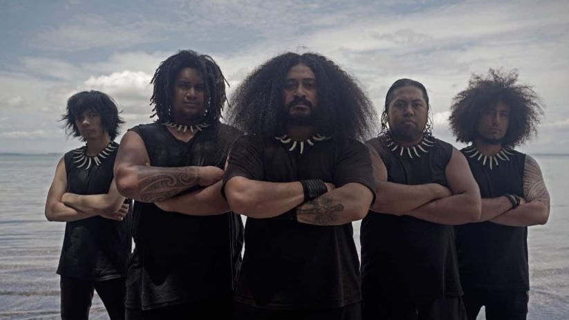 Five men dressed in black and Samoan necklaces cross arms staring at camera with ocean behind them. 