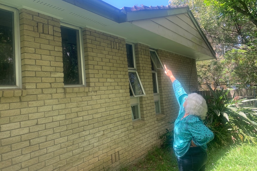 A photo of a woman wearing bright turquoise shirt, grey hair, de-identified, points to a roof of a cream brick house.