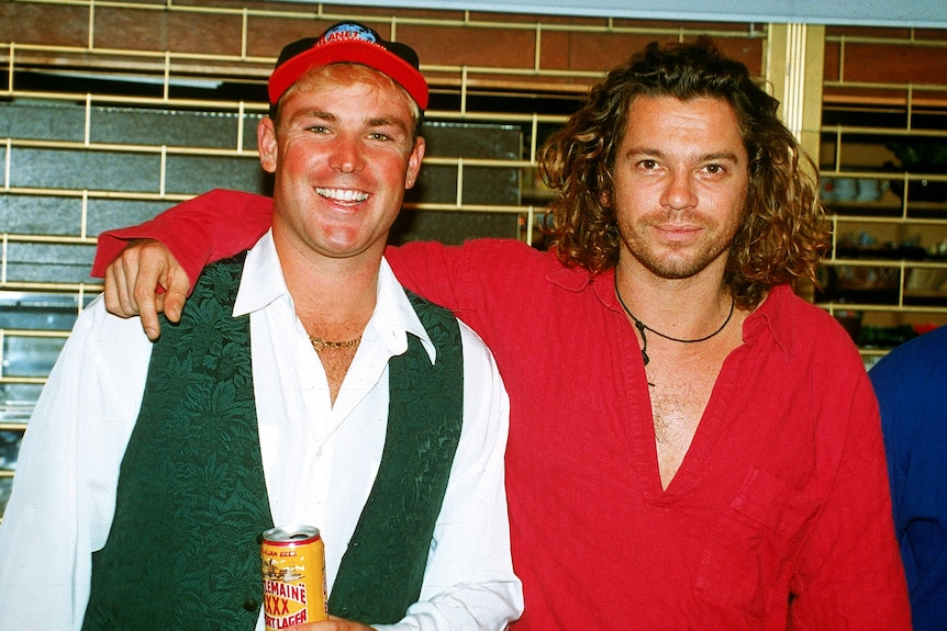 A man in a red shirt with long hair puts his arm around another man in a baseball cap and green vest