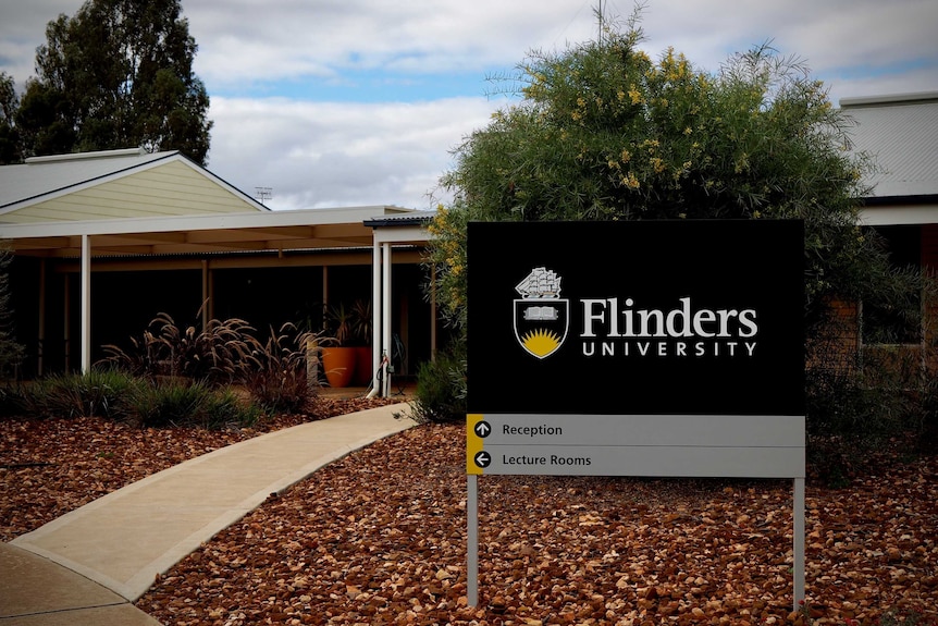 A dark sign sits in front of a yellow brick University building. It reads 'Flinders University'.