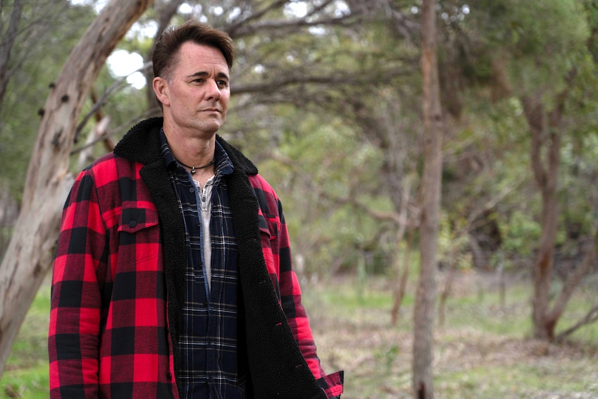 Jarrod wears a tartan jacket and a serious expression while pictured in a natural setting
