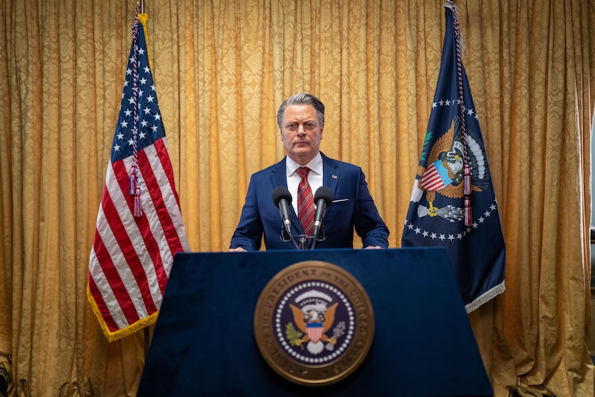 A middle aged white man stands behind a Presidential podium in a suit with red tie. A US and Presidential flag are 