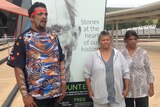 Three Indigenous people stand in front of a sign