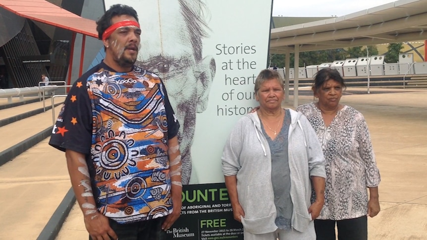 Three Indigenous people stand in front of a sign