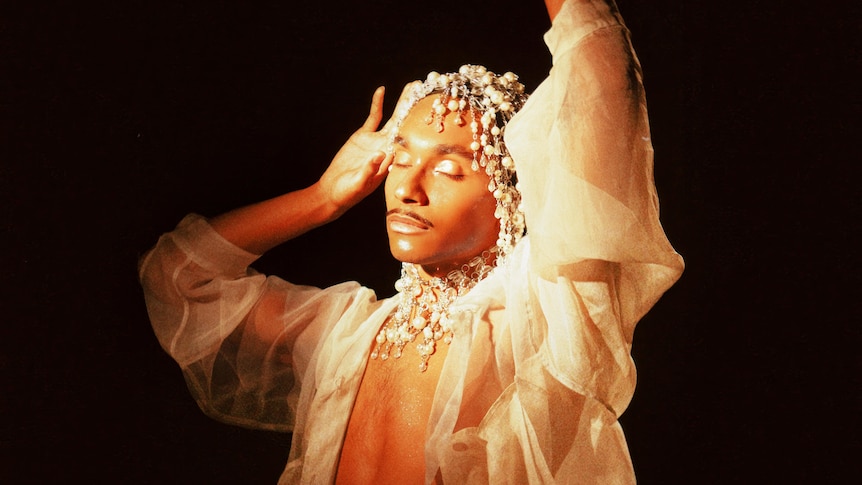 Forest Claudette wears a see-through blouse, make-up and pearl headpiece against black backdrop