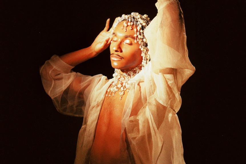 Forest Claudette wears a see-through blouse, make-up and pearl headpiece against black backdrop