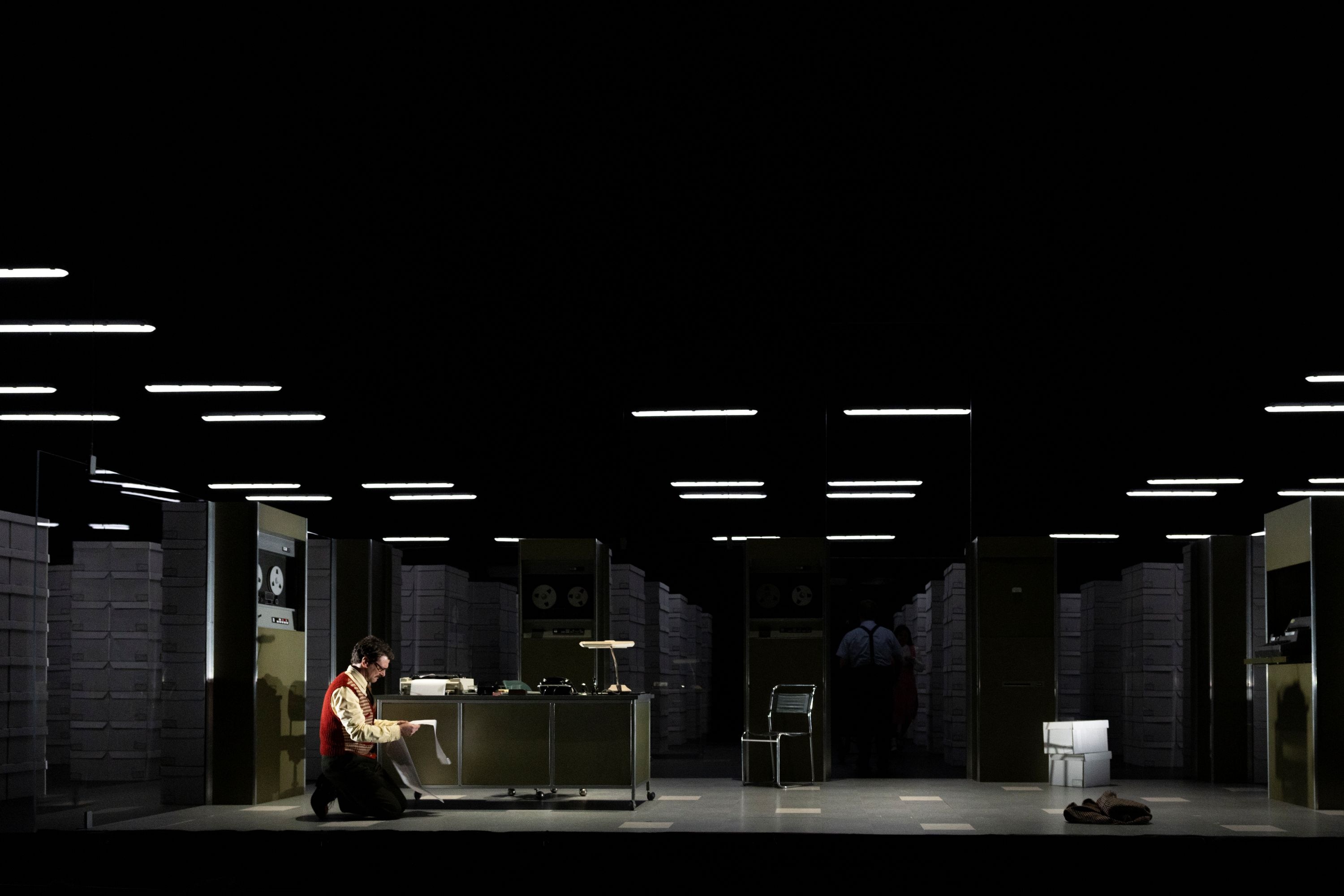 On stage, Dan Spielman, 47, in glasses, kneels on the ground in front of a desk and chair, looking over pages of code.
