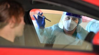 A person in bioprotective gear including mask, stands at a passenger side window holding a swab test.