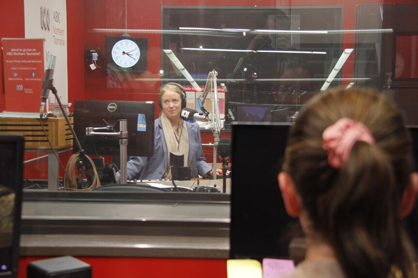 A producer looks through a glass screen to a woman presenting radio in an ABC studio. 