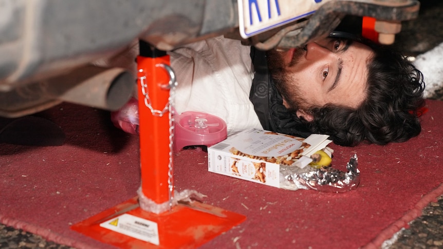 A long-haired, bearded man lying underneath a car.