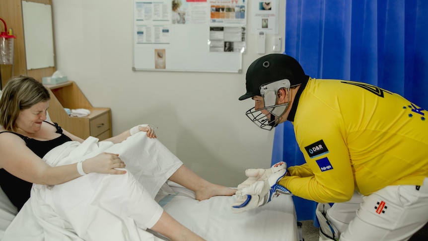 A man wearing cricket uniform, wicketkeeping gloves and helmet in the hospital with his pregnant wife for a unique photoshoot.  