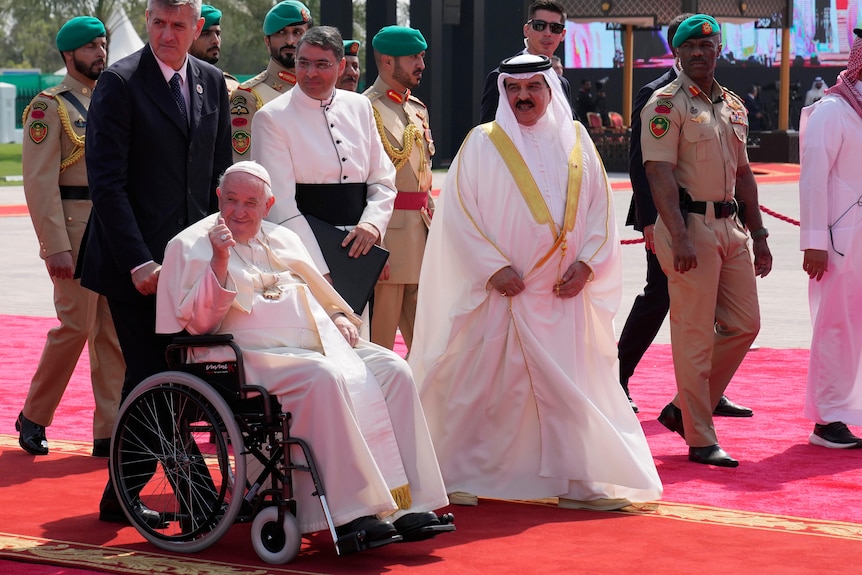 Pope Francis gives a thumbs up from a wheelchair surrounded by staff, military and Bahrain's king. 