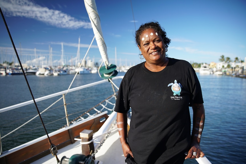 A woman with traditional white markings on her face, wearing a black t-shirt, stands on a ship, smiling.