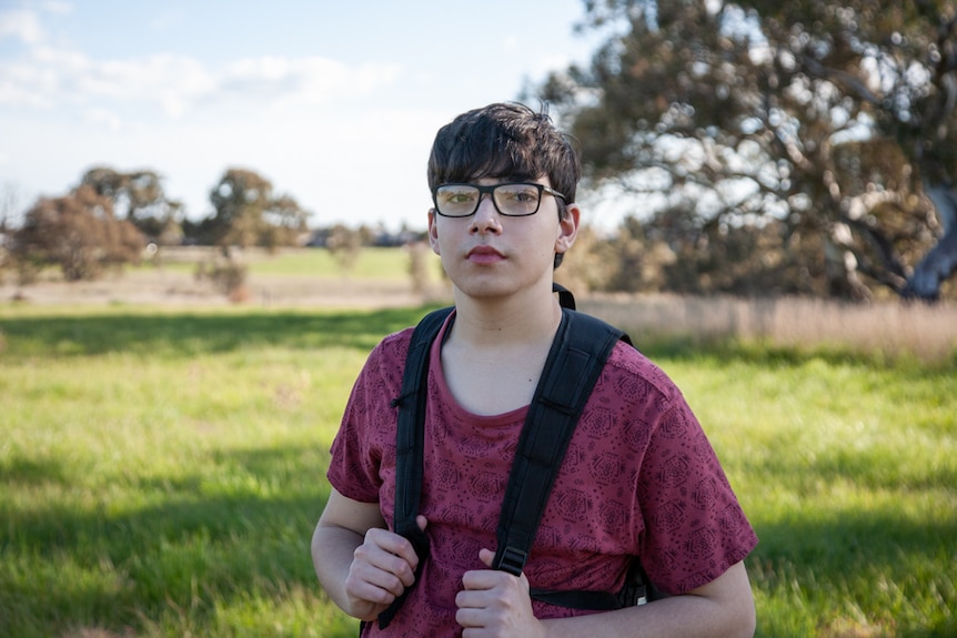 A boy in a red shirt and glasses grips the straps of his backpack as he looks at the camera