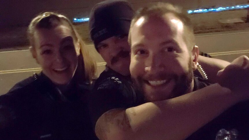 A police officer grabs another man around the neck and a female police officer stands behind. All three are smiling.